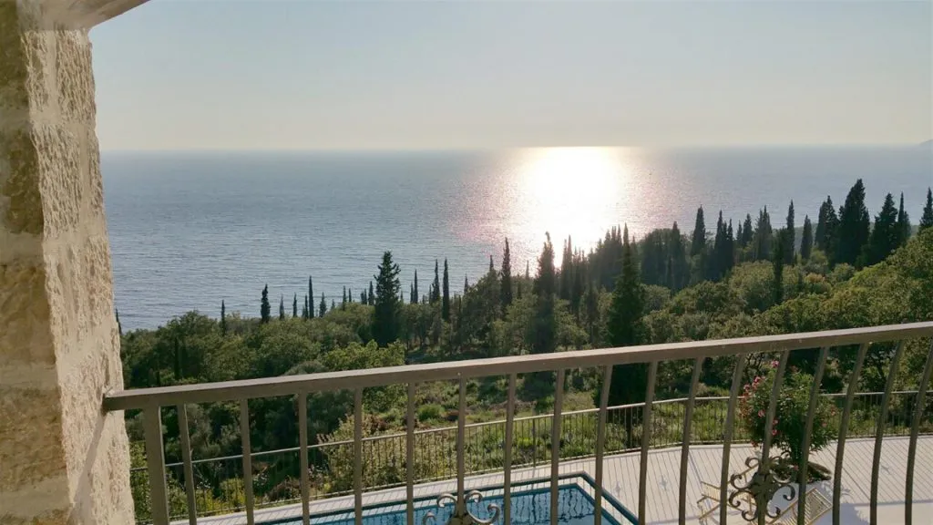 View from a balcony overlooking the sea and forest. A stone wall, metal railing, and part of a pool are visible in the foreground.