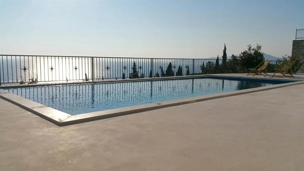 Outdoor swimming pool with sea view, surrounded by a metal fence and wooden deck chairs.