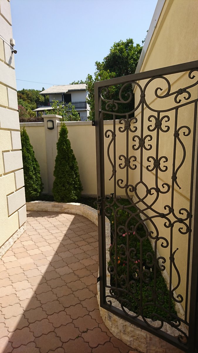 Wrought-iron gates and a well-kept courtyard with thuja trees at a villa in Utjeha, Montenegro. Tiled paving and landscape design.