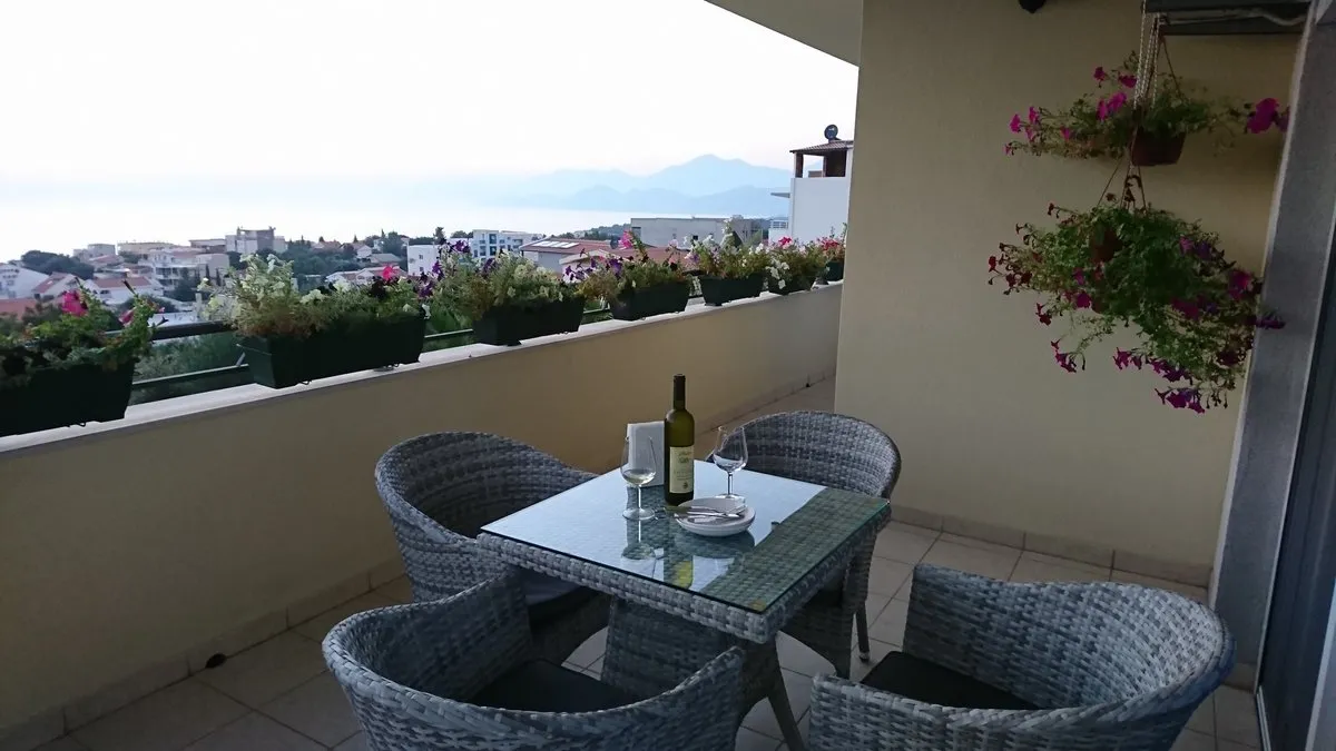 Balcony with wicker furniture, wine bottle, and flower boxes on the railing overlooking a coastal town.