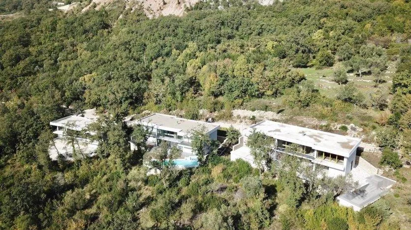 Aerial view of a modern white house nestled in a dense green forest with a visible swimming pool.