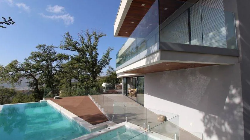Modern house with a swimming pool, wooden deck, glass railings, and surrounding trees under a blue sky.
