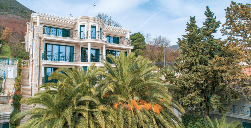 Three-story villa with stone cladding, wrought-iron balconies, and palm trees in Bijela, Montenegro. Front view of the building.