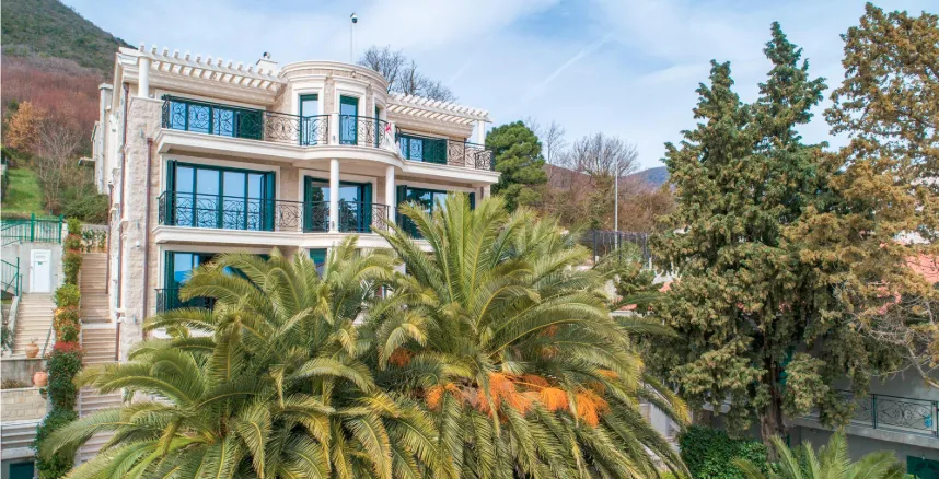 Exterior view of a multi-story villa with balconies and palm trees against mountains.
