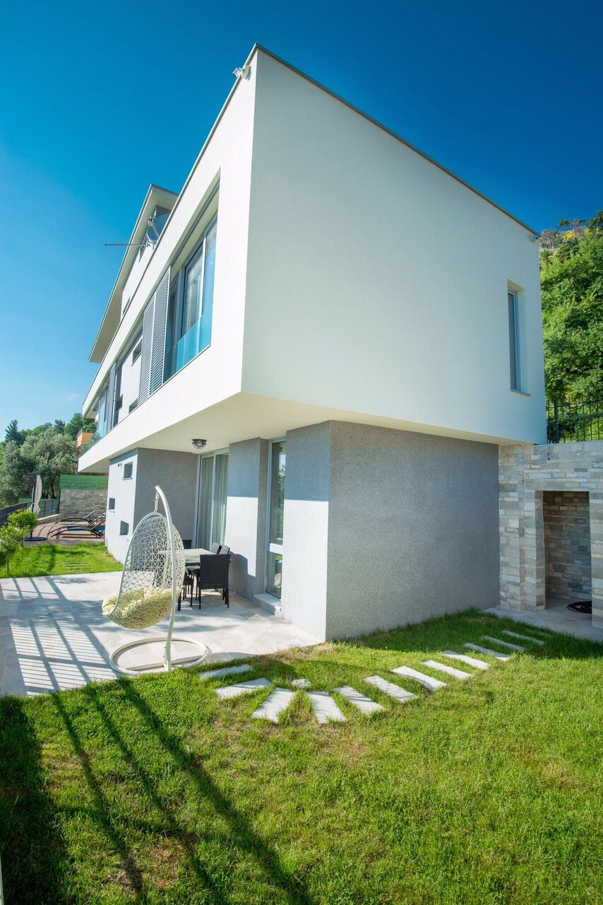 Modern villa in Kotor, Montenegro: view of the terrace with a hanging chair, garden, and the building facade with panoramic windows.