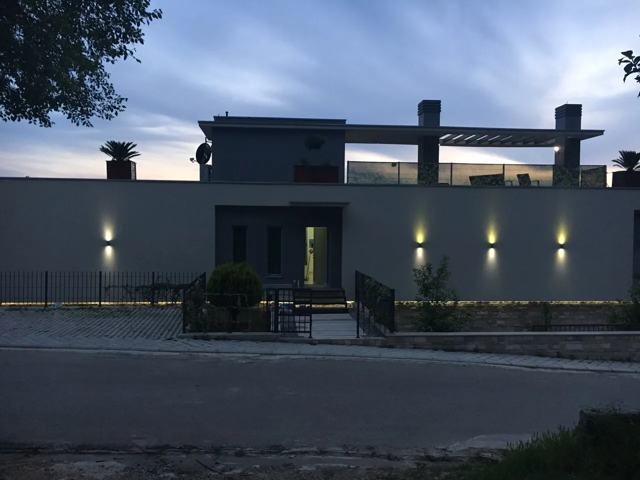 Exterior view of a modern two-story house with a terrace and wall lighting at dusk.