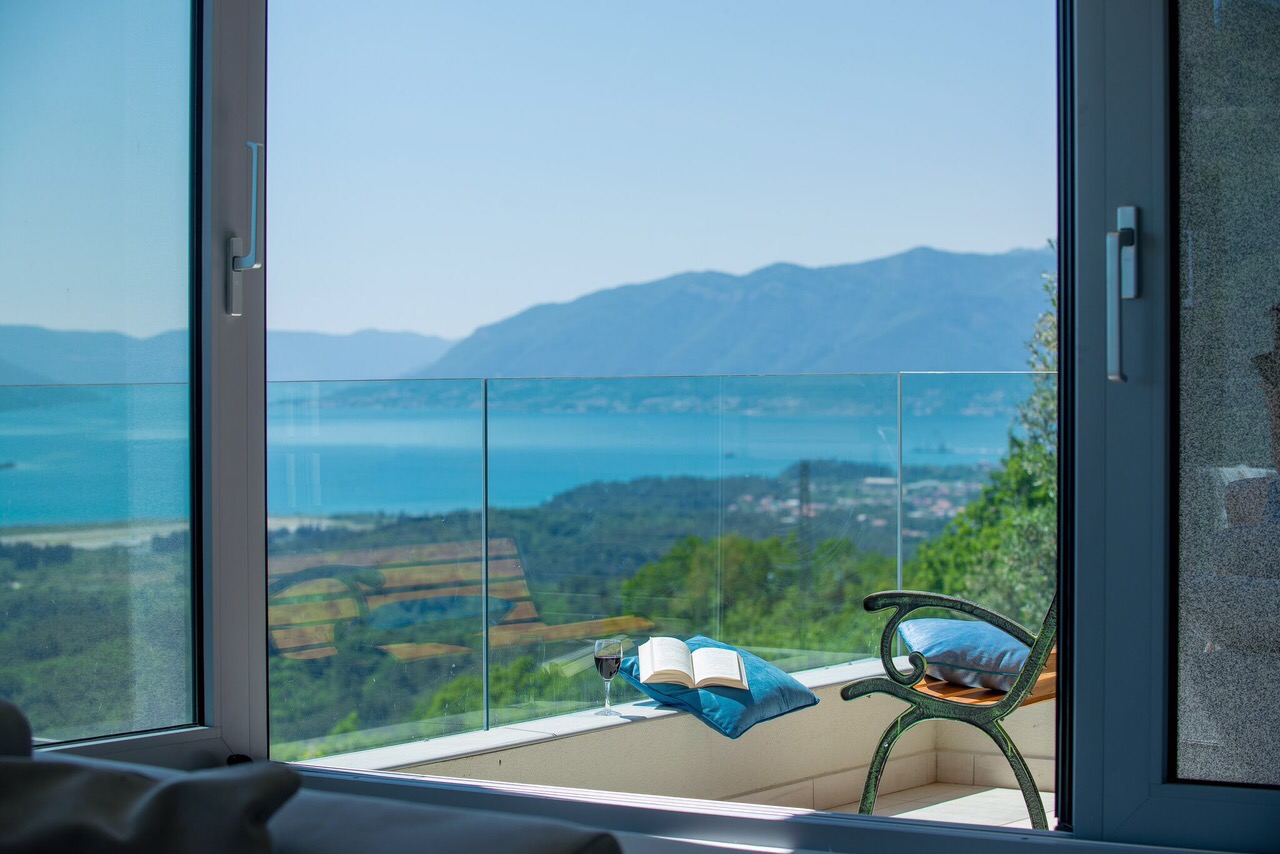 View from the villa balcony in Kotor, Montenegro: glass railing, chair, book, and panoramic view of the Bay of Kotor.