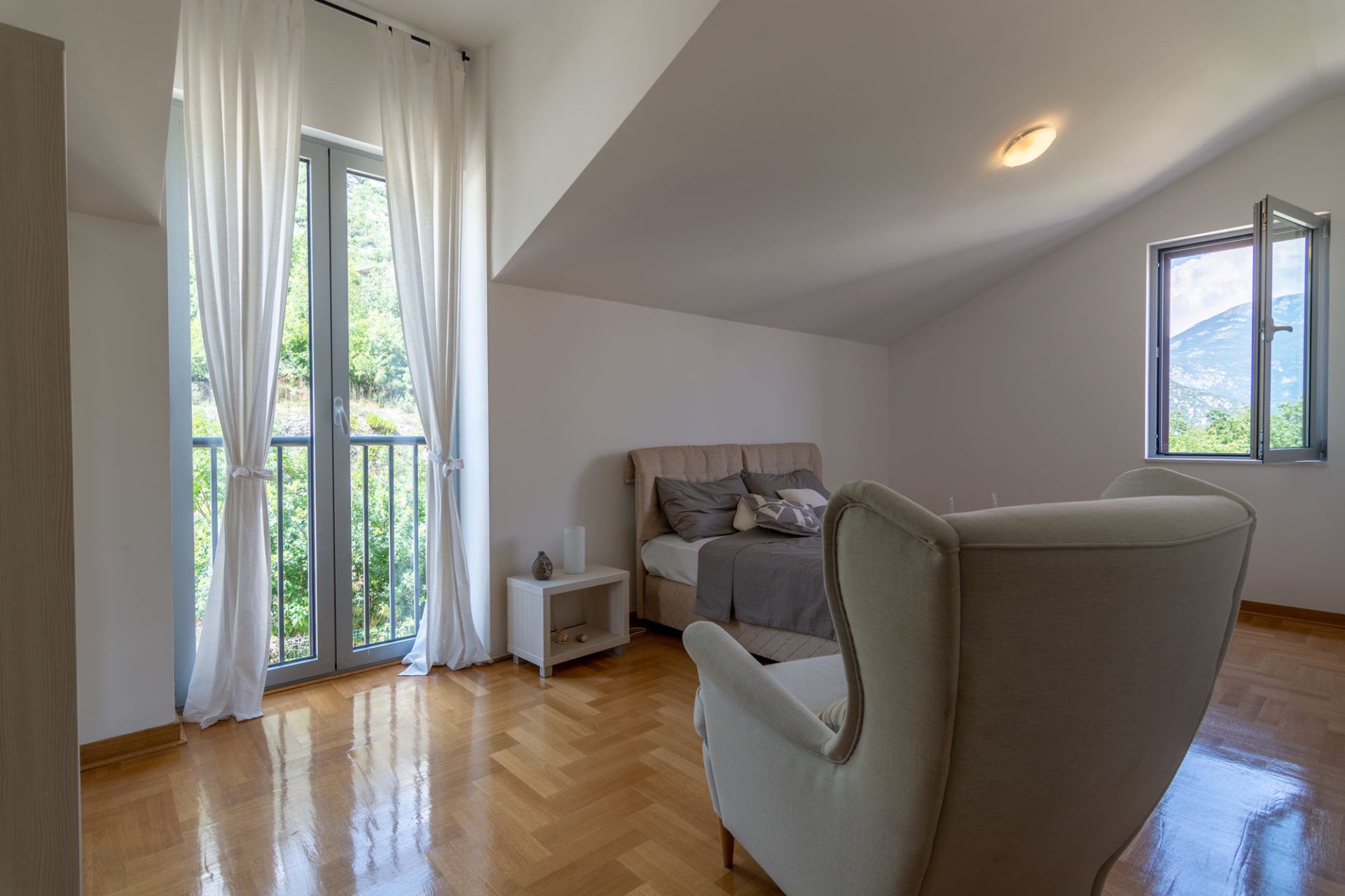 Bright bedroom in an apartment in Morinj, Montenegro: a double bed, an armchair, parquet flooring, and a mountain view from the window.