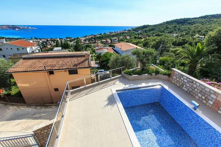 Open-air swimming pool with sea and mountain views by the house in Šušanj, Montenegro, with a terrace and glass railing.