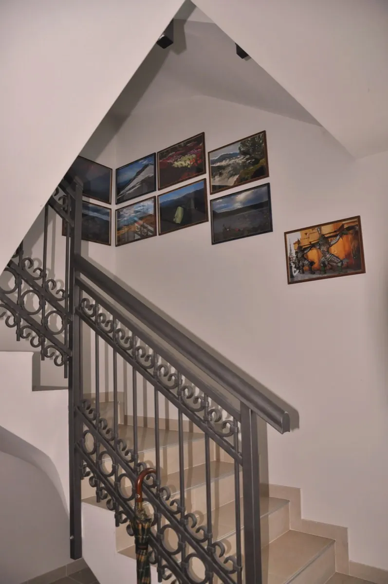 Staircase with ornate wrought iron railing and a collection of landscape paintings on the white wall below the ceiling.
