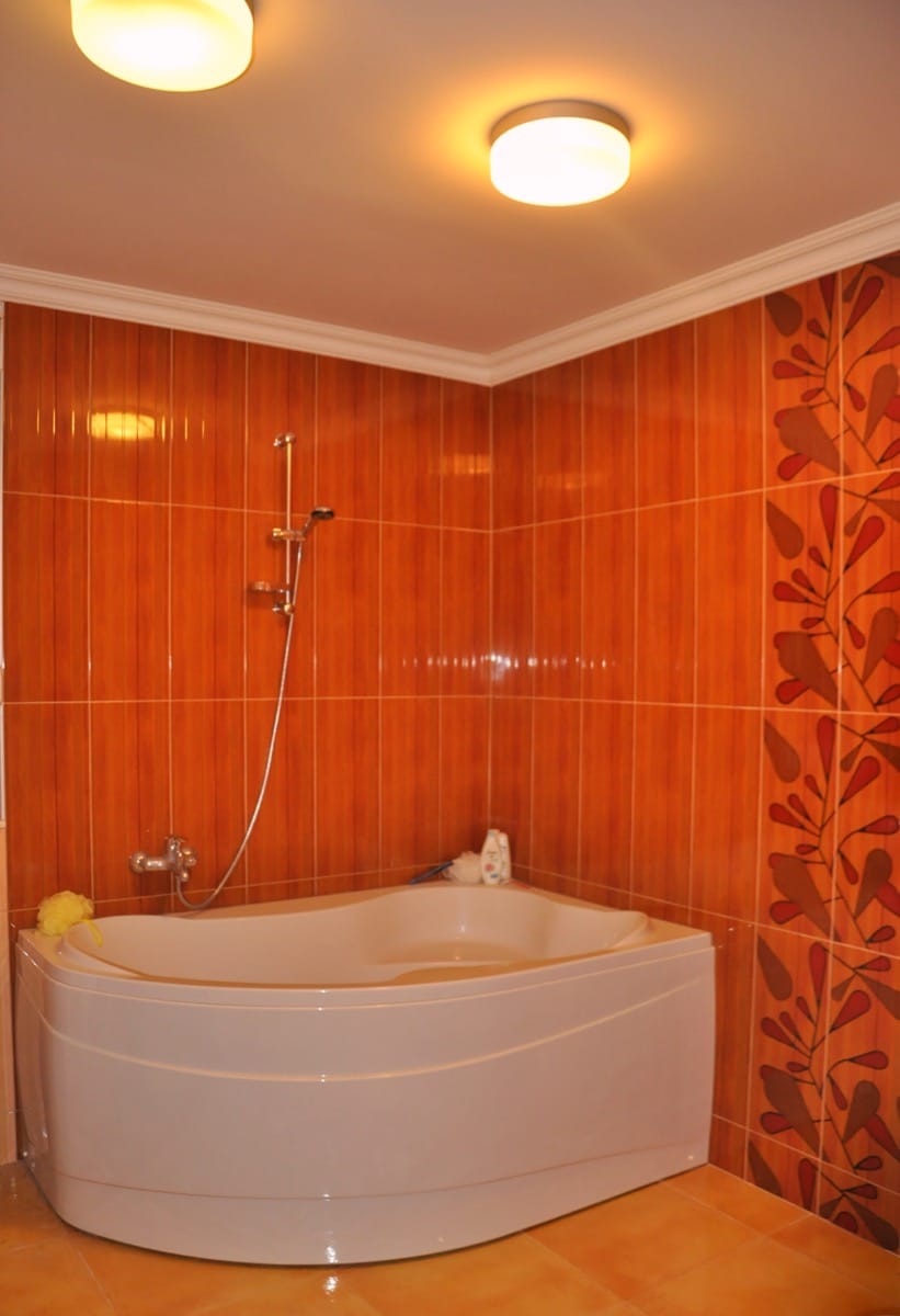 White corner bathtub with a shower rail against orange tiled walls featuring a leaf pattern.