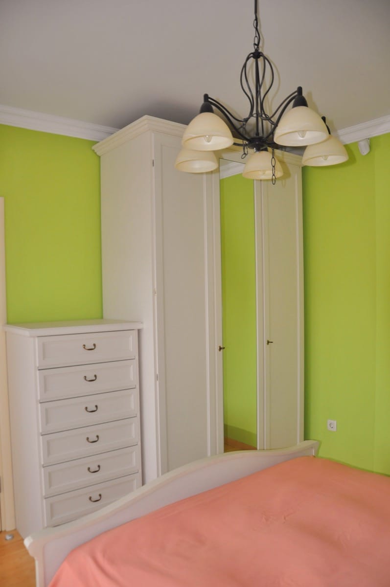 Bright bedroom with lime green walls, white wardrobe, chest of drawers, and a bed with pink bedding visible.