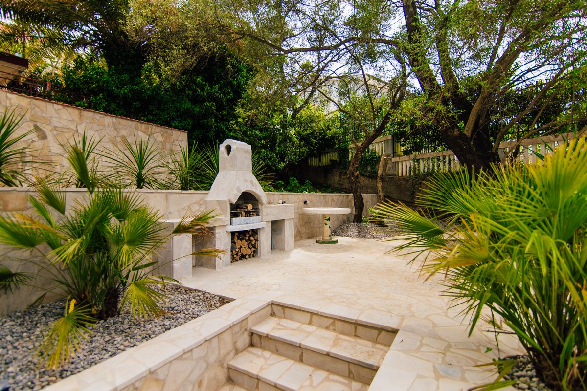 Barbecue area with a stone grill and patio on the grounds of a villa in Krašići, Montenegro, surrounded by palm trees and trees.