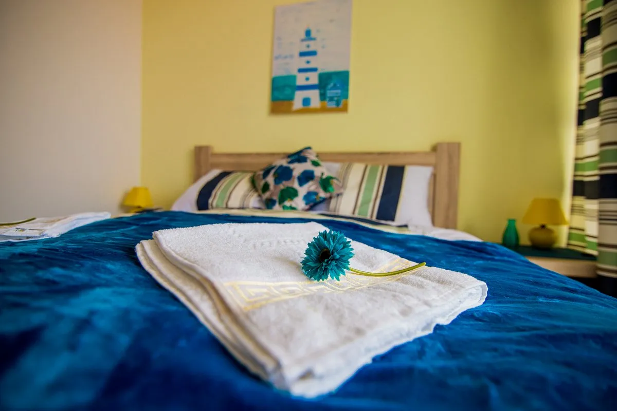 Bed with blue cover, white towels and blue flower against a yellow wall with lighthouse art.