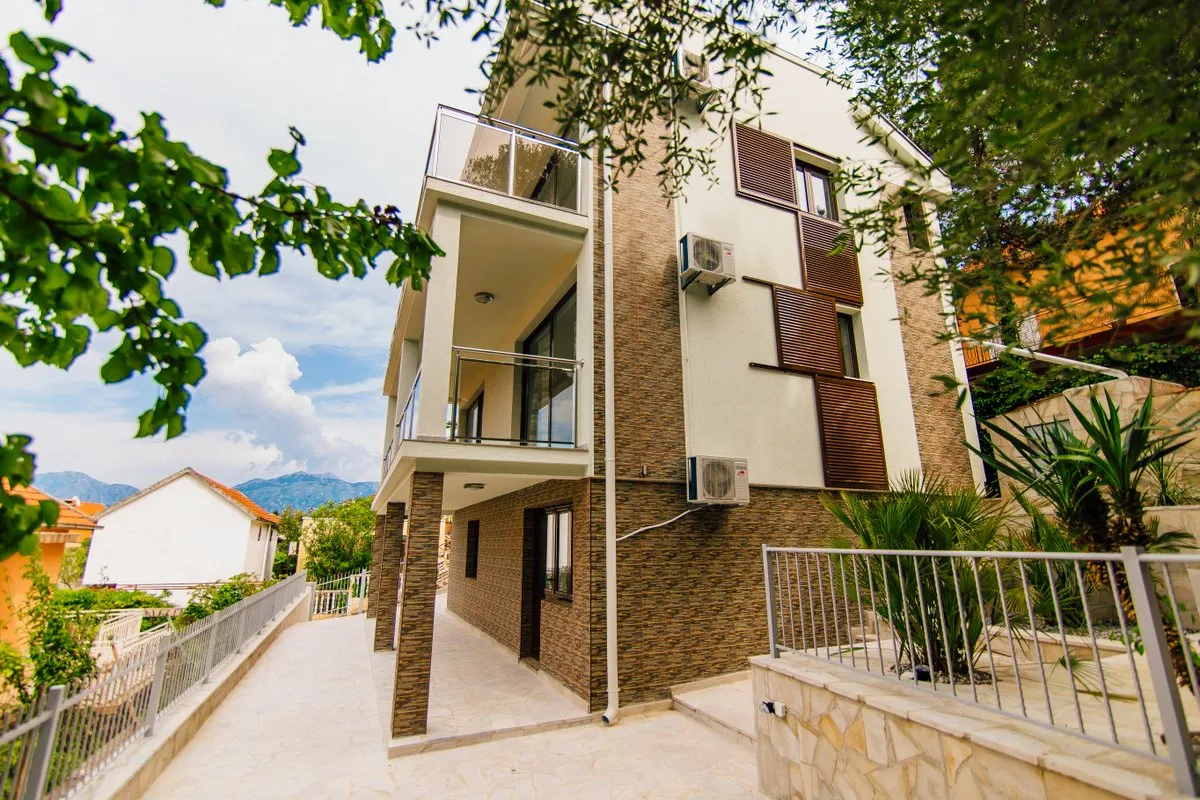 Exterior view of a modern multi-story house with balconies, stone cladding, and AC units against a mountain backdrop.