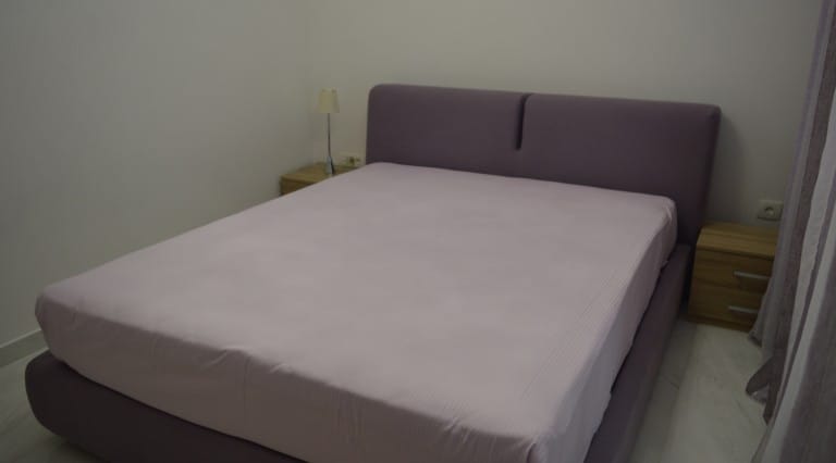 Bedroom featuring a purple double bed, wooden nightstands, and a small lamp against white walls.