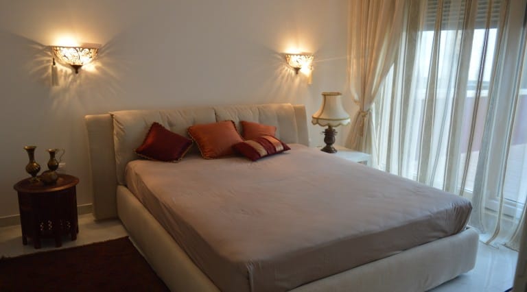 Bedroom featuring a large bed with decorative pillows, wall sconces, and sheer curtains by the window.