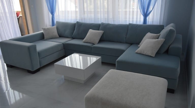 Blue L-shaped sofa with gray pillows and a white glossy coffee table on light tiled flooring.
