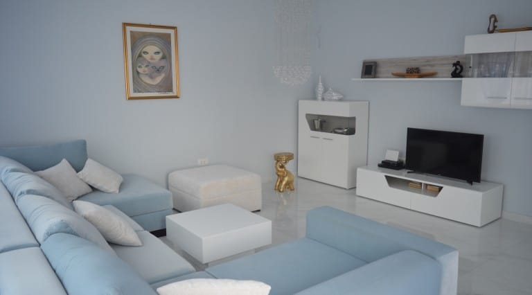 Bright living room in an apartment in Ljuta, Montenegro: blue corner sofa, white furniture, TV area, and marble floor.