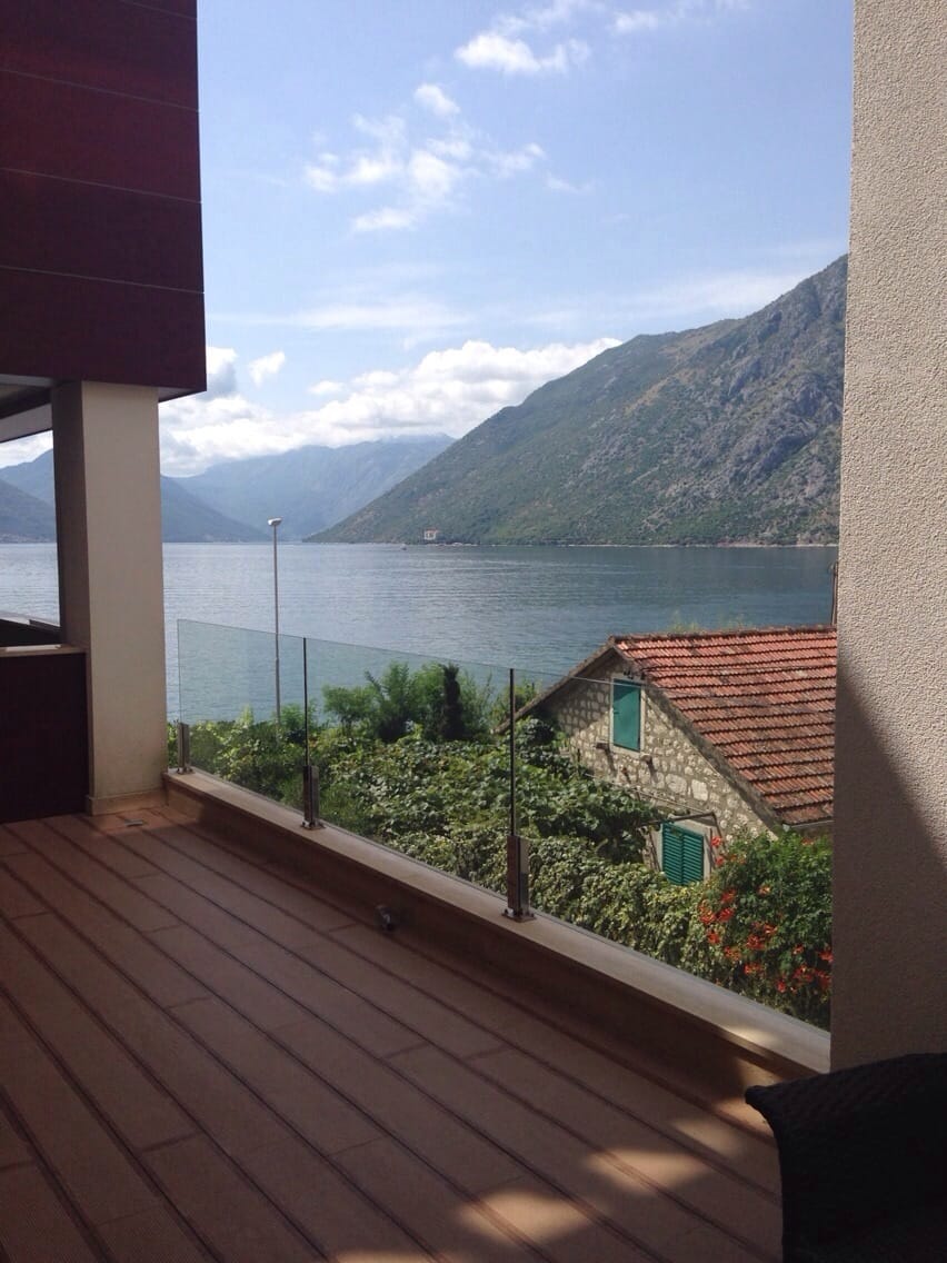Wooden balcony with glass railing overlooking the sea and mountains.