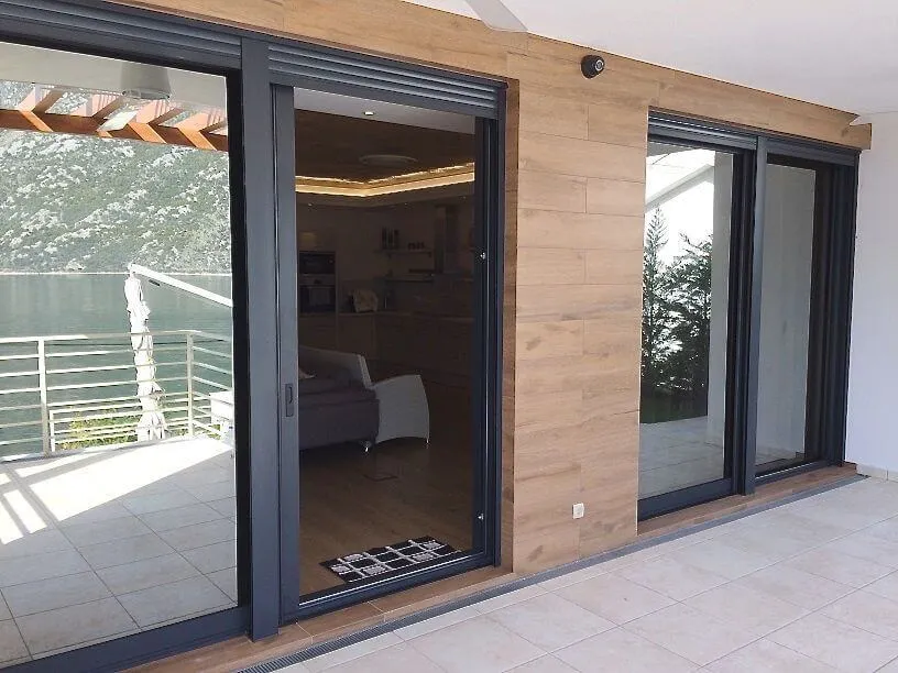 Tiled terrace with water view, large sliding glass doors, wood-paneled wall and interior living space visible.
