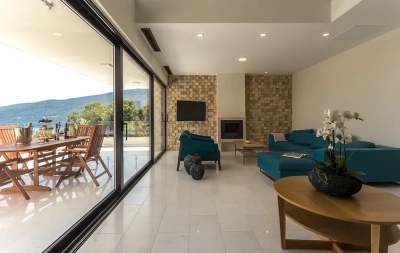 Bright living room with a blue sofa, stone fireplace, and TV. Large glass sliding doors open to a terrace overlooking mountains and sea.