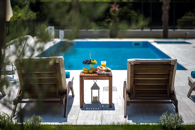Wooden loungers and a table with fruit by the edge of a blue swimming pool on a sunny terrace.