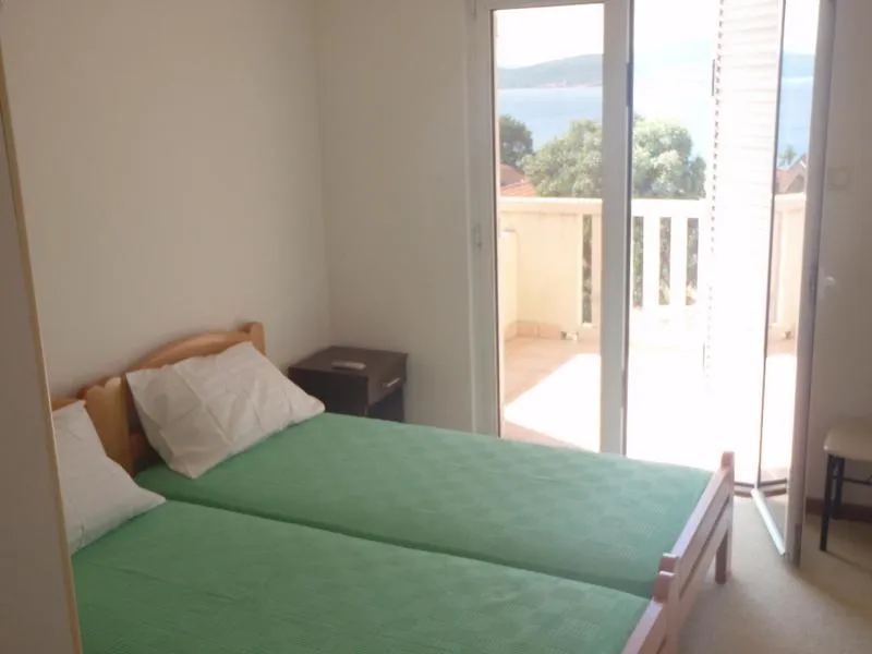 Two single beds with green covers, a nightstand, and an open door leading to a balcony overlooking water.
