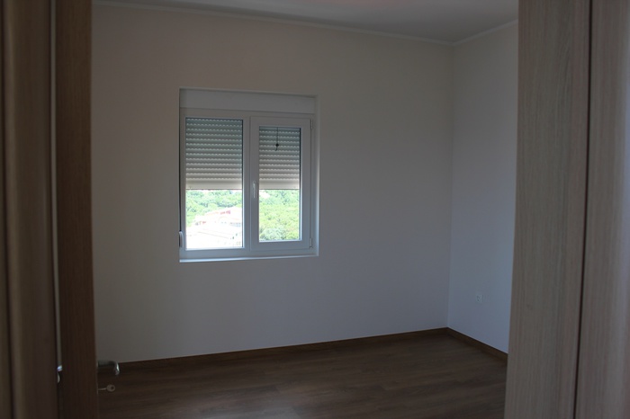 Bright bedroom in an apartment in Dobra Voda, Montenegro: laminate flooring, white walls, a window with roller shutters, and a view of greenery.
