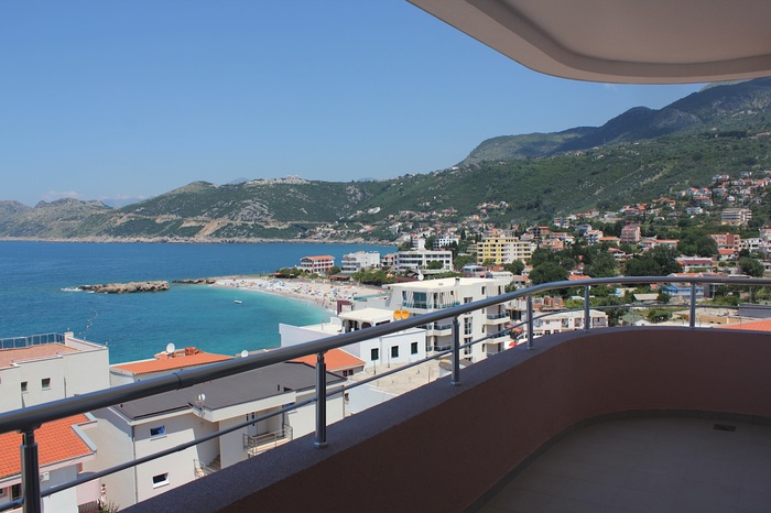 View from the apartment balcony in Dobra Voda, Montenegro: a panorama of the sea, beach, and mountains with modern terrace finishes.