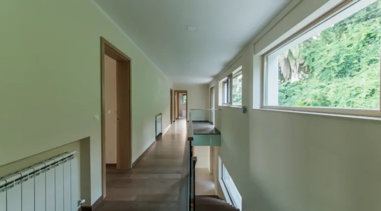 Long hallway with wooden floors, white walls, a radiator, and a glass railing overlooking a lower level.