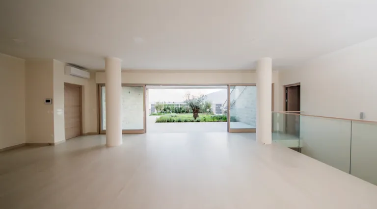 Empty bright room with white columns and glass railing, overlooking a garden through sliding doors.
