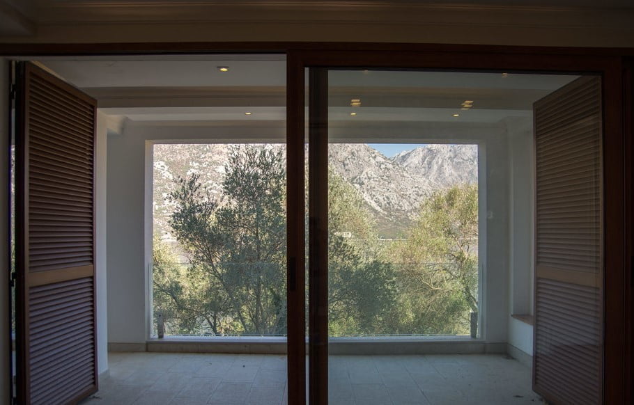 View from the villa balcony in Stoliv, Montenegro: panoramic windows, wooden shutters, and a landscape with mountains and olive trees.