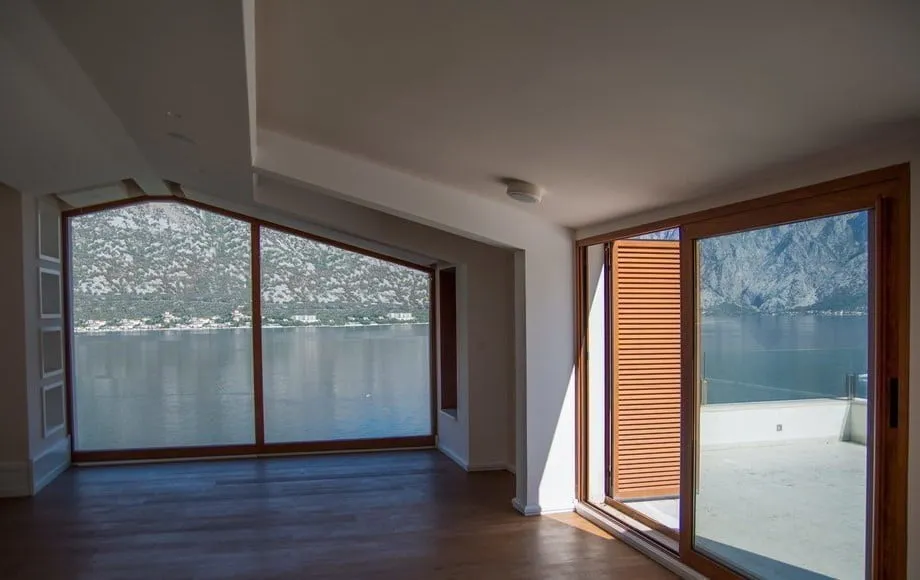 Empty room with wooden flooring and large windows offering a view of the water and mountains.