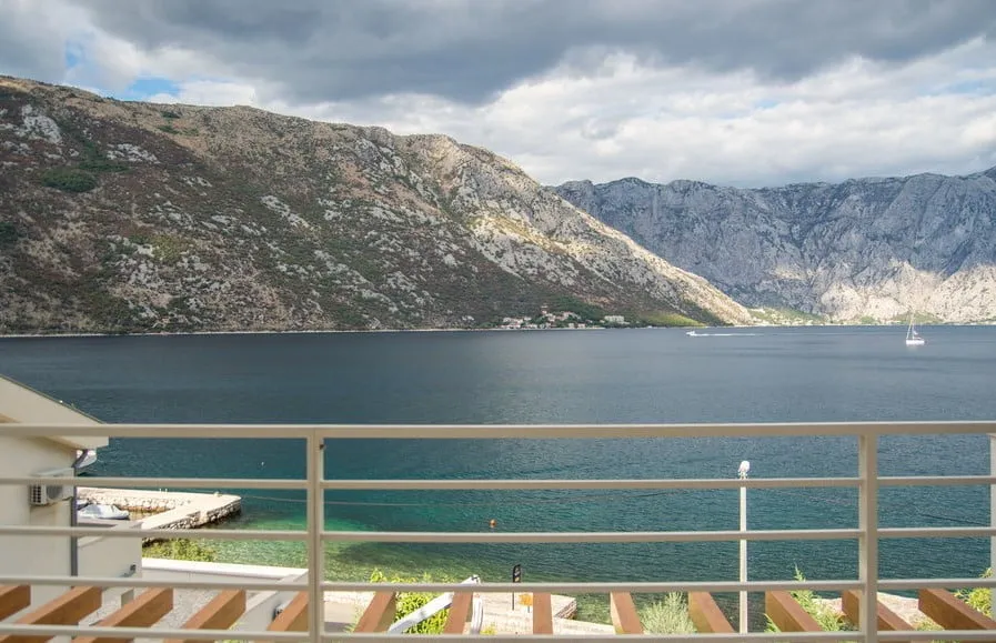 View from a balcony overlooking the sea and mountains. A white railing and terrace part are visible.