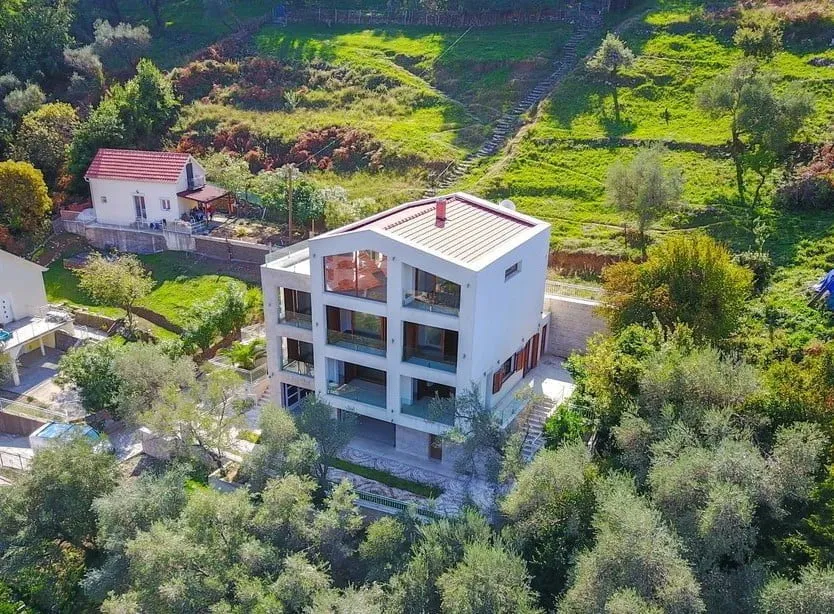 Aerial view of a modern white multi-story house with balconies, situated on a green hillside surrounded by trees.