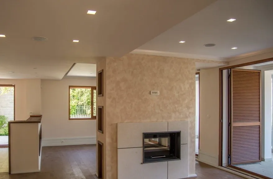 Bright room featuring a central fireplace, wooden flooring, and an open layout connecting to a kitchen area.