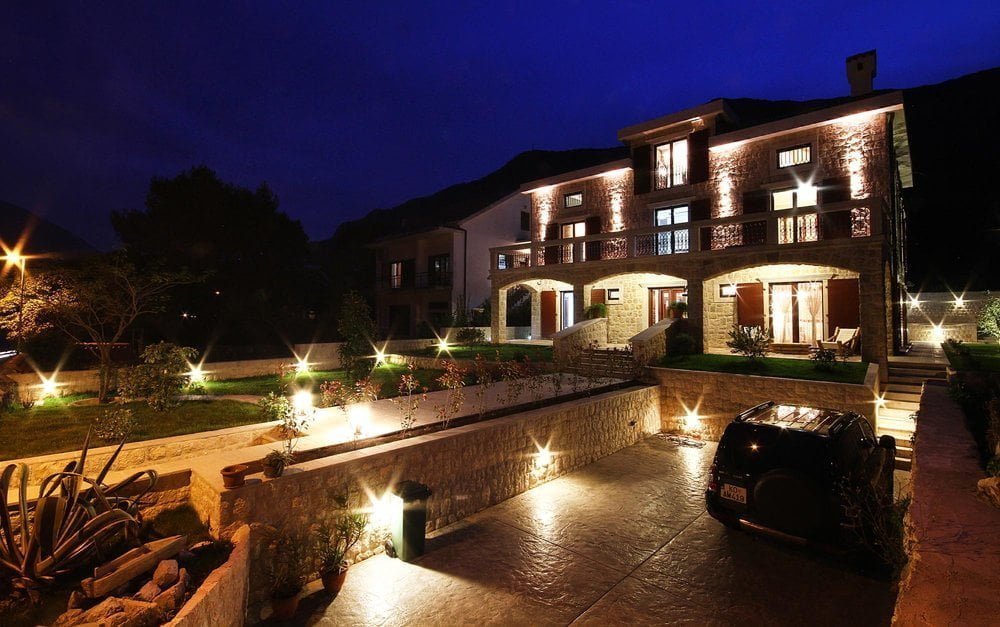 Two-story stone villa with facade lighting, terrace, and parking in Prčanj, Montenegro, night view.