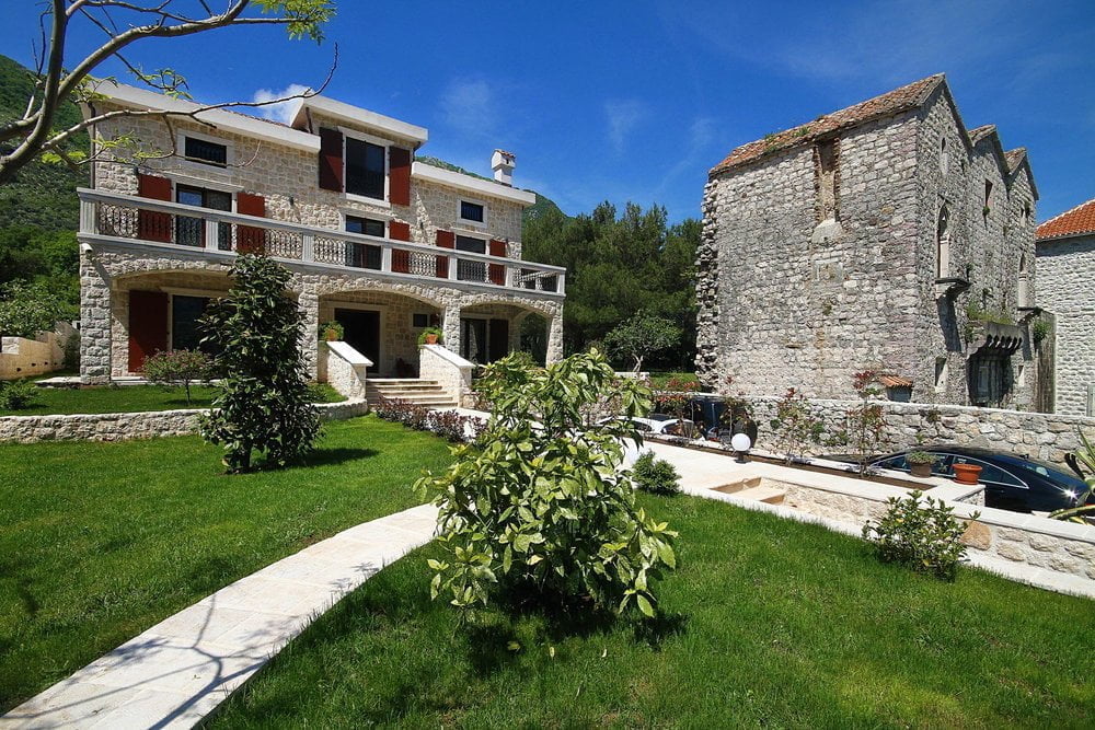 Two-story stone villa with a terrace and a green garden in Prčanj, Montenegro, against the backdrop of a historic stone building.