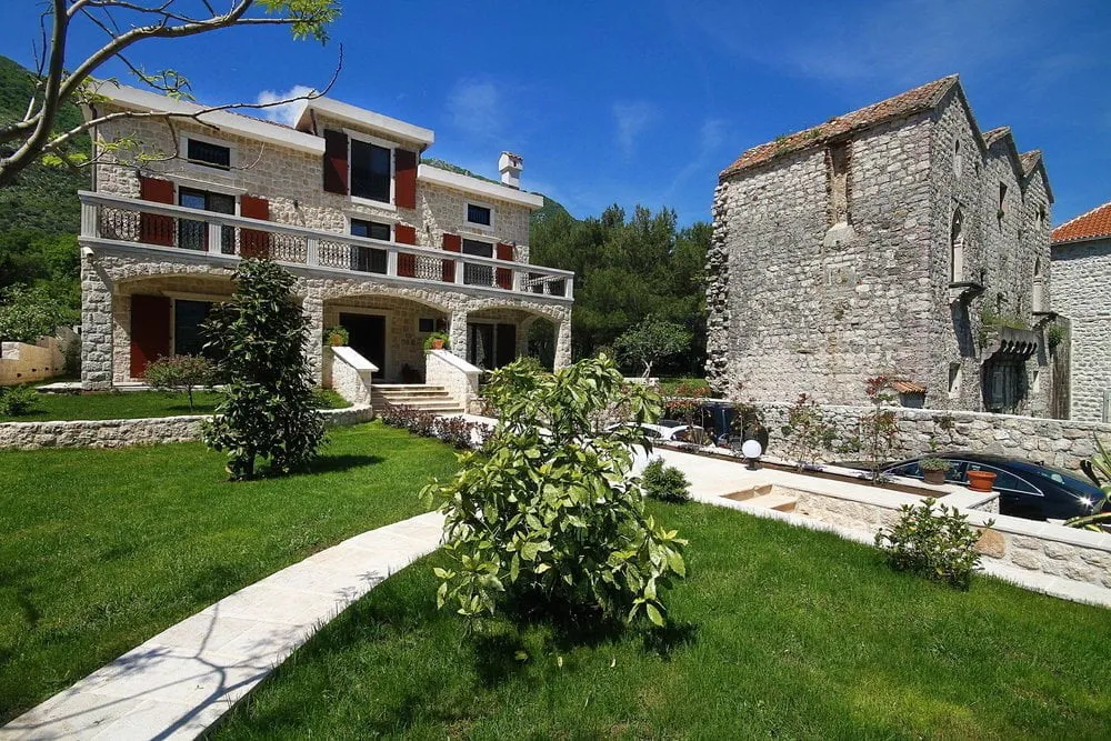 Stone house with balcony and red shutters, green lawn, pathway, and old stone building under blue sky.