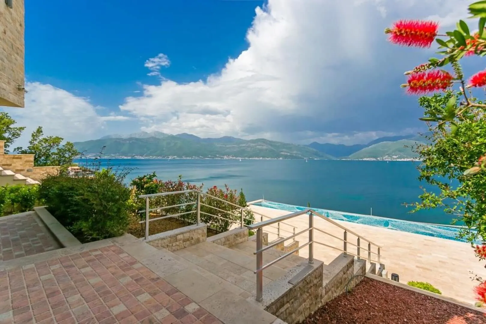 Infinity pool terrace overlooking the sea and mountains with stone masonry and red flowers.
