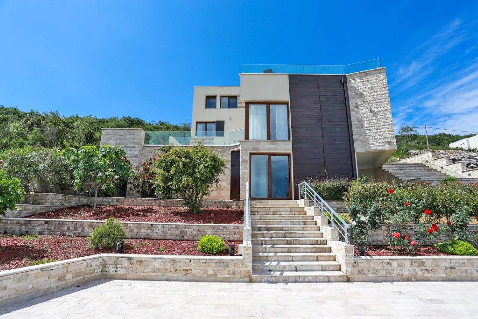Modern villa in Krašići, Montenegro: facade with stone cladding, terrace, and staircase against a green garden backdrop.