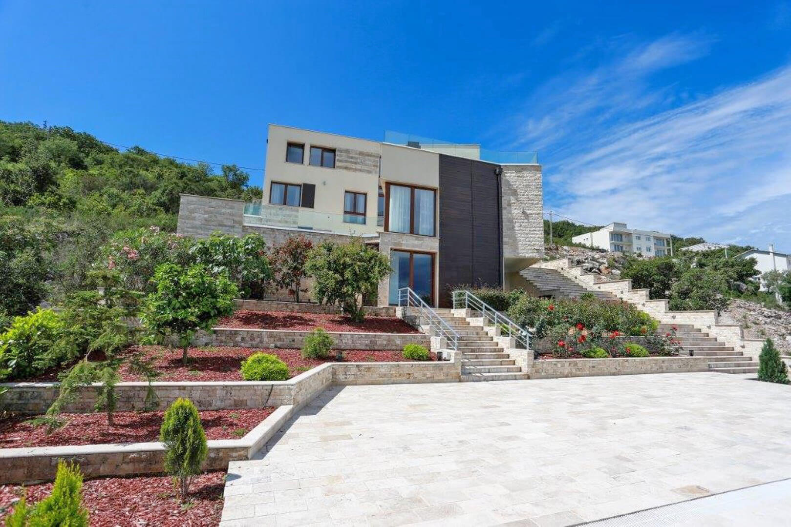 Modern villa in Krašići, Montenegro: stone-clad facade, terraced garden, and a wide staircase against a mountain backdrop.