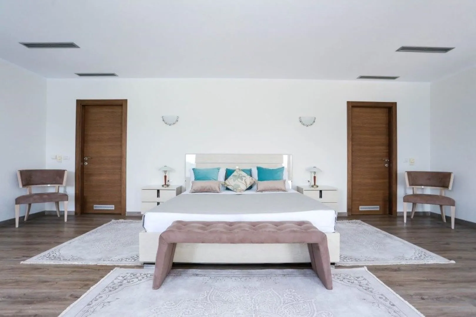 Bright bedroom featuring a double bed with turquoise pillows, wooden doors, side tables, and patterned rugs on wood flooring.