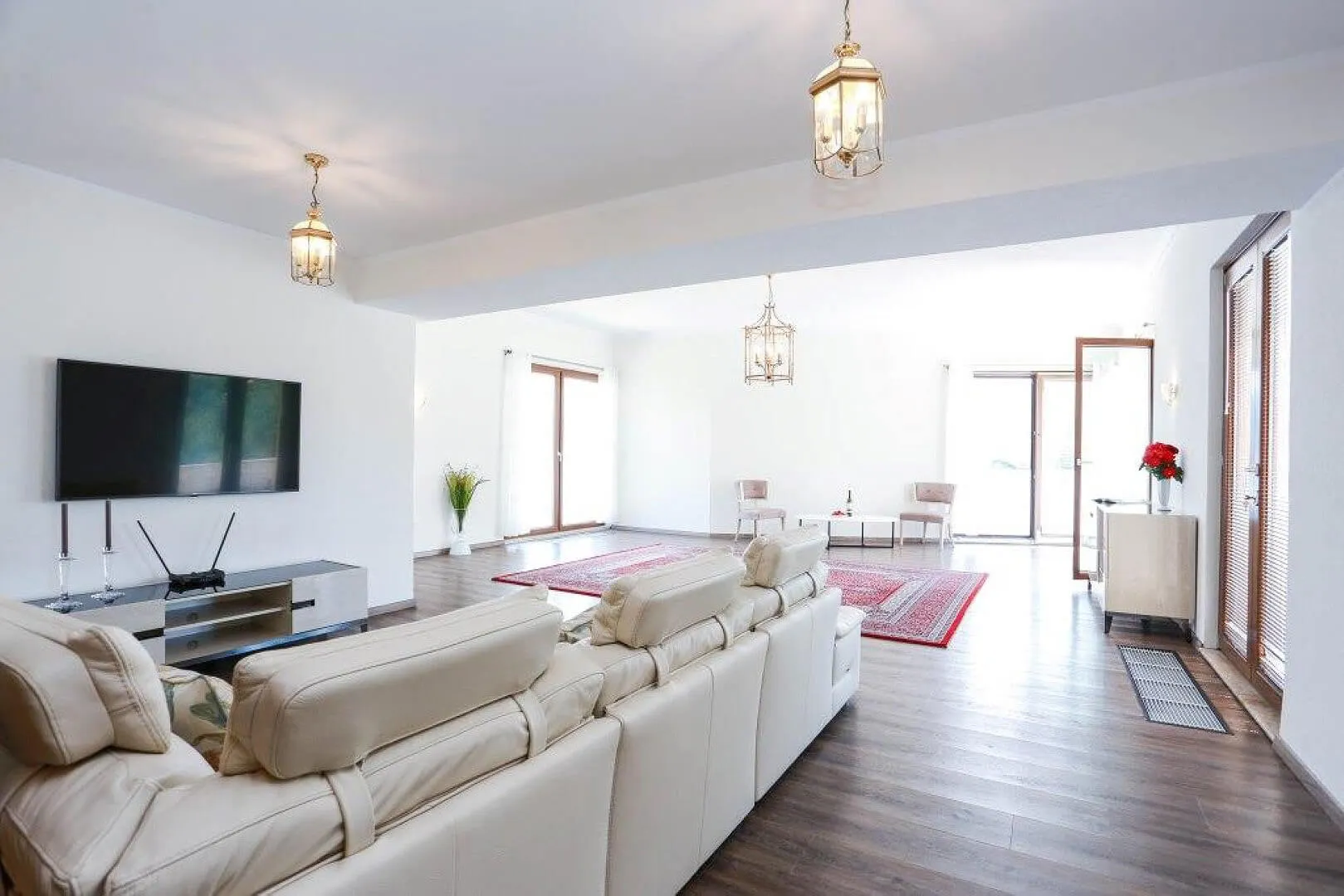 Bright living room featuring a white leather sofa, wall-mounted TV, red rug, and hanging lantern-style lights.