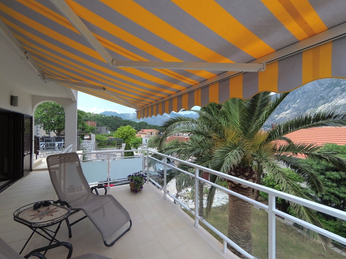 Villa balcony in Prčanj, Montenegro: striped awning, sun lounger, small table, and panoramic view of the mountains and palm trees.