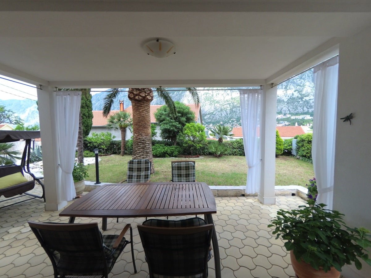 Covered terrace of a villa in Prčanj, Montenegro: dining area, view of the garden, palm trees and mountains, garden furniture and swing.