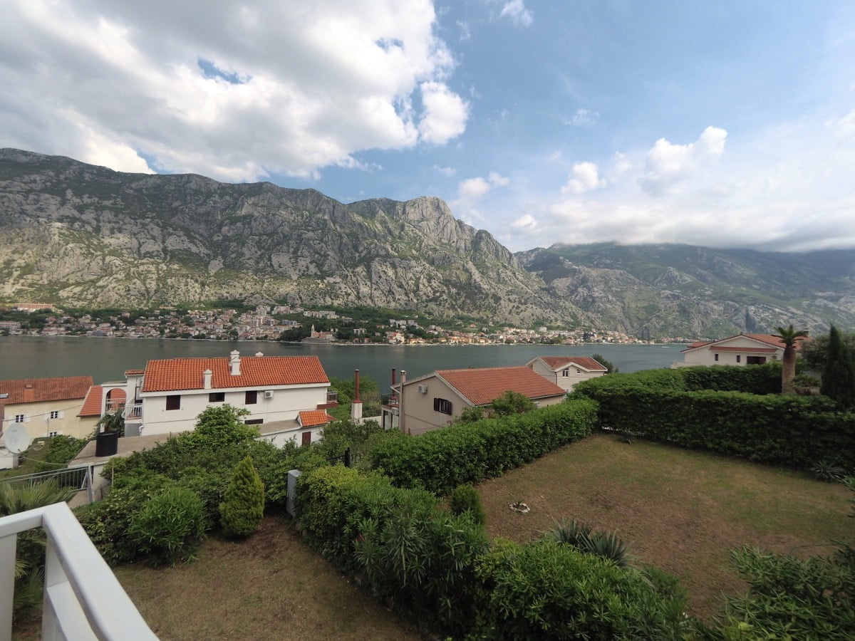 View from the villa balcony in Prčanj, Montenegro: a panorama of the Bay of Kotor, mountains, and a well-kept green garden.