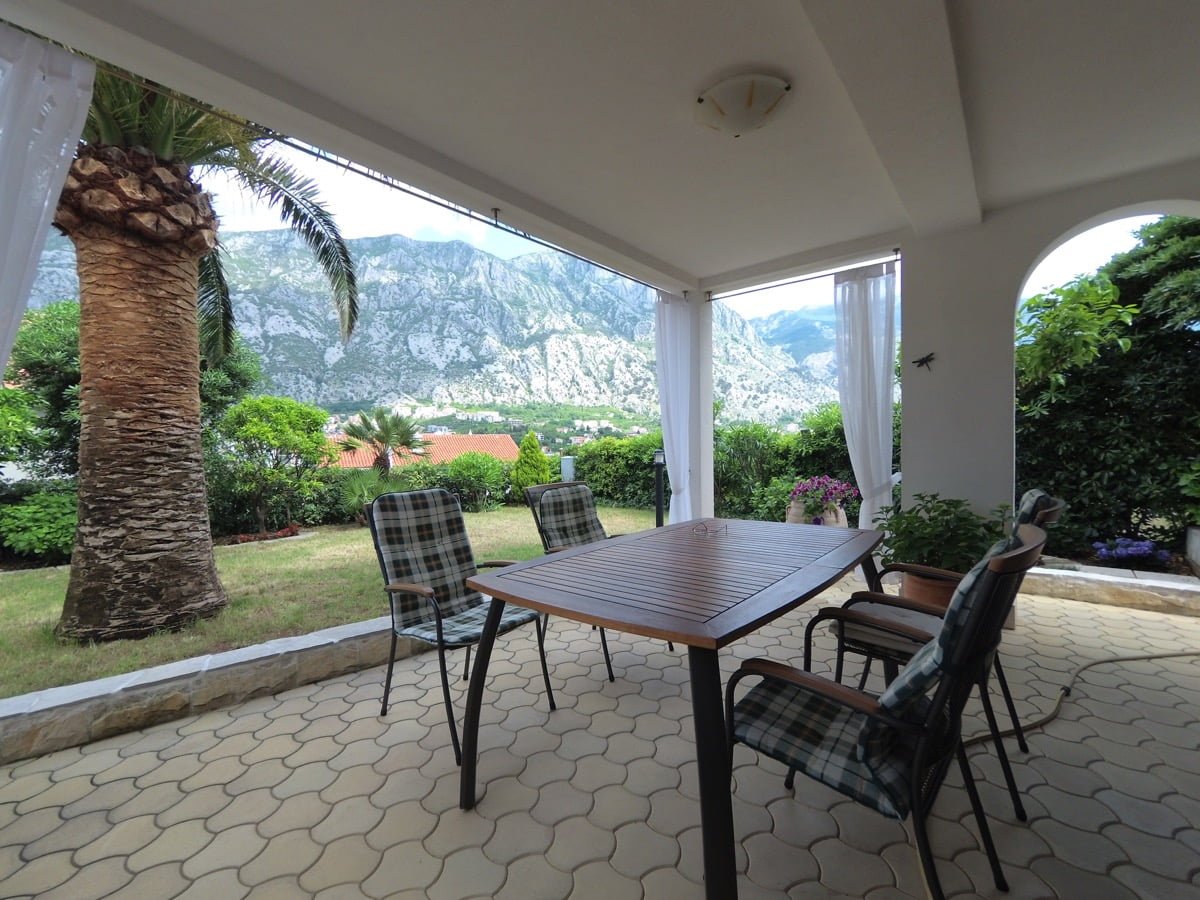 Covered terrace of a villa in Prčanj, Montenegro: dining area, mountain view, garden with a palm tree, and paved area.