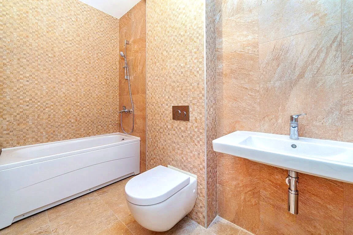 Bathroom featuring a white bathtub, wall-mounted toilet, and sink set against beige mosaic tiles.